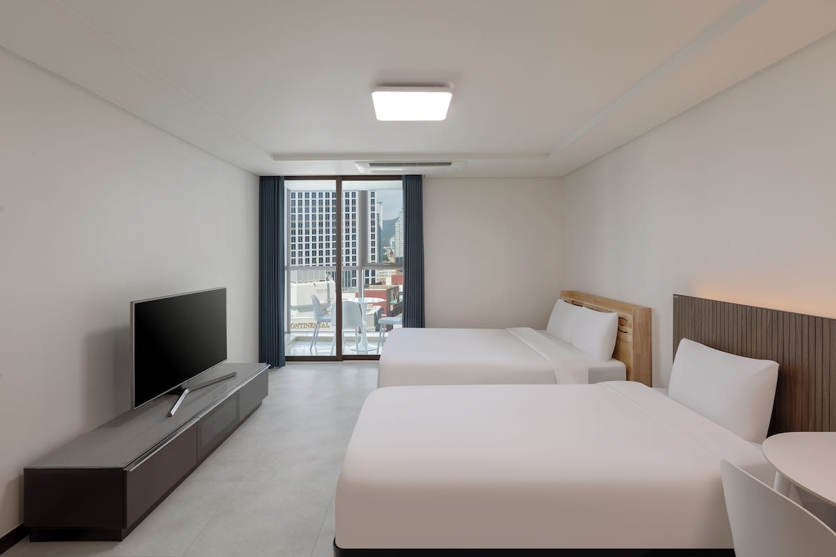 Habitación de hotel moderna con una televisión de pantalla plana, dos camas, una mesita y vistas a la ciudad a través de amplias ventanas.