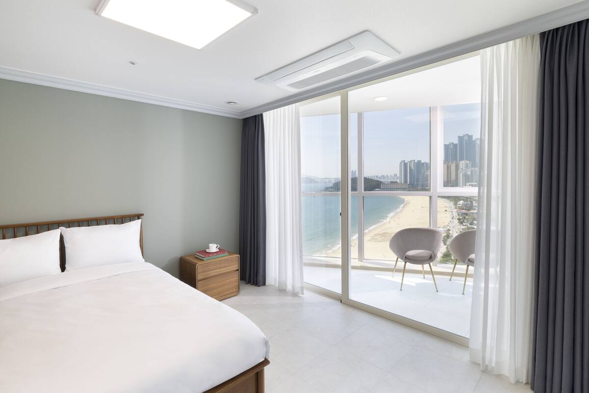 Habitación de hotel con una cama grande, una mesita de noche, vista a la playa y al paisaje urbano.