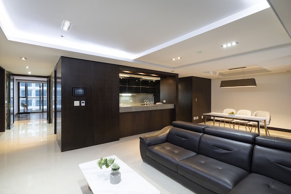 A modern living room with a dark leather sofa, a white coffee table with a plant, and a kitchen area with a built-in island and seating.