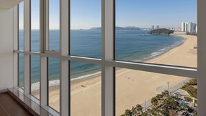 A view from a high-rise window overlooking a beach with a clear sky, sandy shore, and distant buildings. - Pale De CZ (Busan)