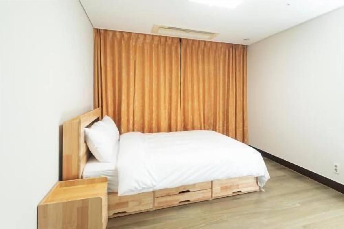 Una cama individual con cabecera y cajones de madera, una mesita de noche de madera y una rejilla de ventilación en el techo.