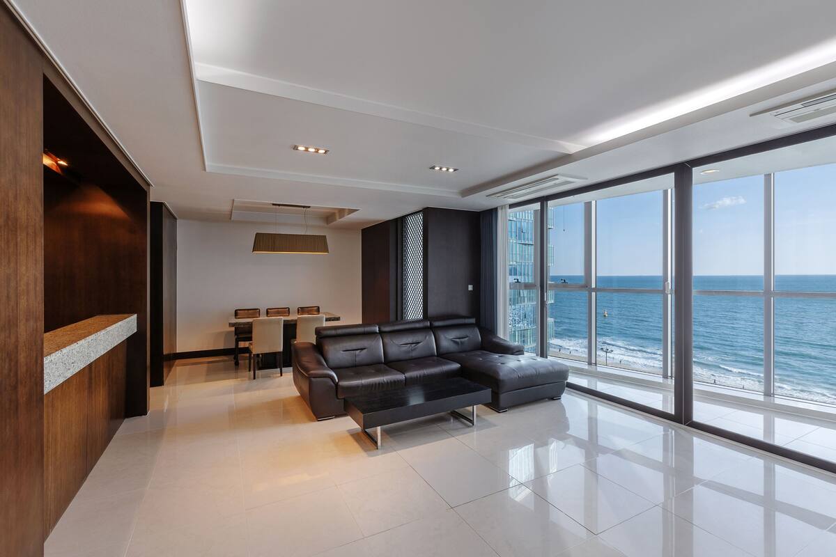 Una sala de estar moderna con un sofá de cuero negro, una mesa de comedor con sillas y ventanas amplias con vistas al mar.