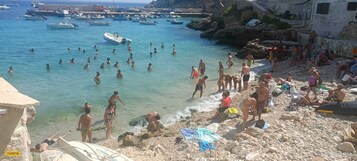 A crowded beach with people swimming and sunbathing, boats in the water, and rocky terrain.