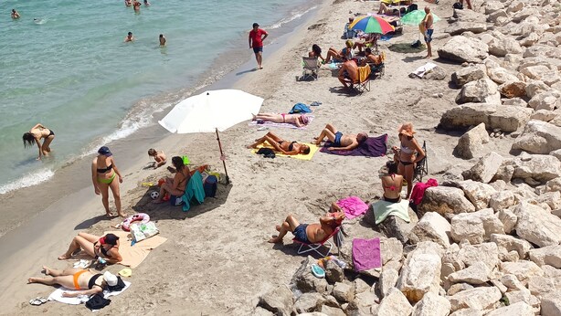 Uma praia com pessoas se banhando ao sol e nadando, um trecho costeiro rochoso e edifícios ao fundo.