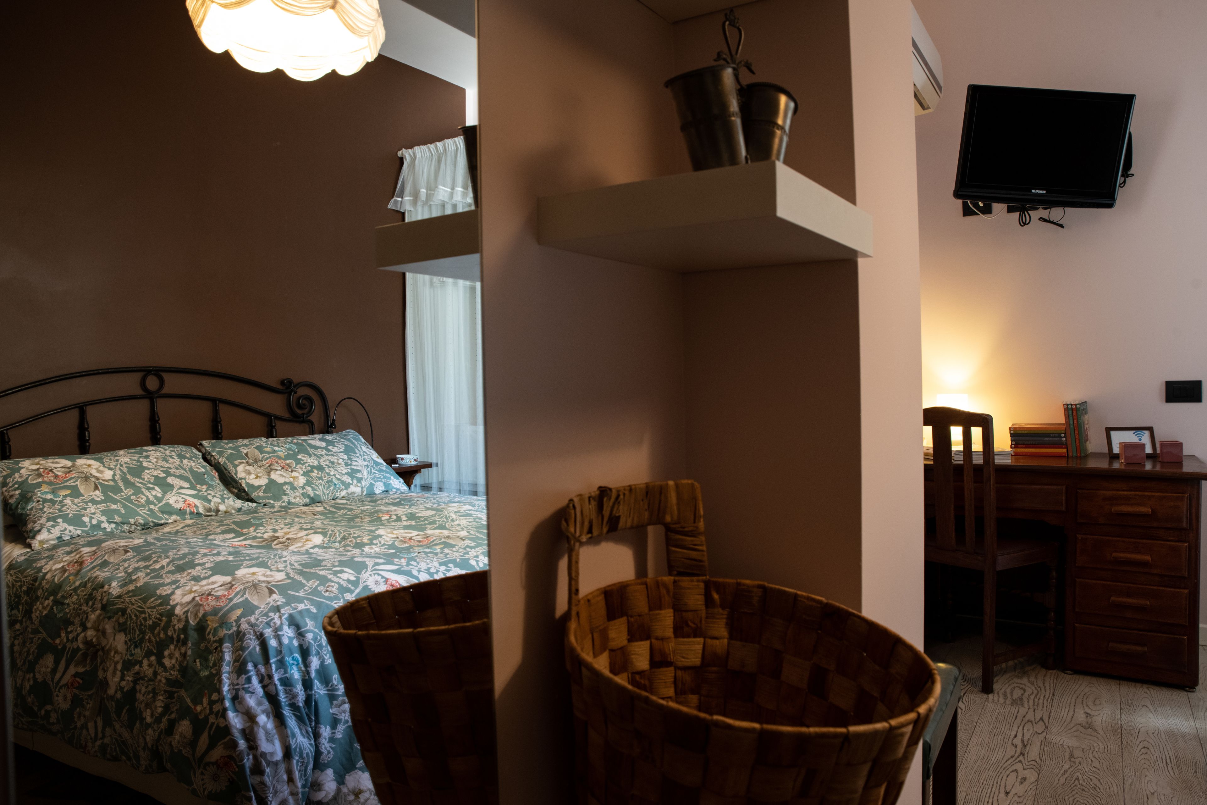 A bedroom with a bed, a wicker chair, a desk, and a television mounted on the wall.