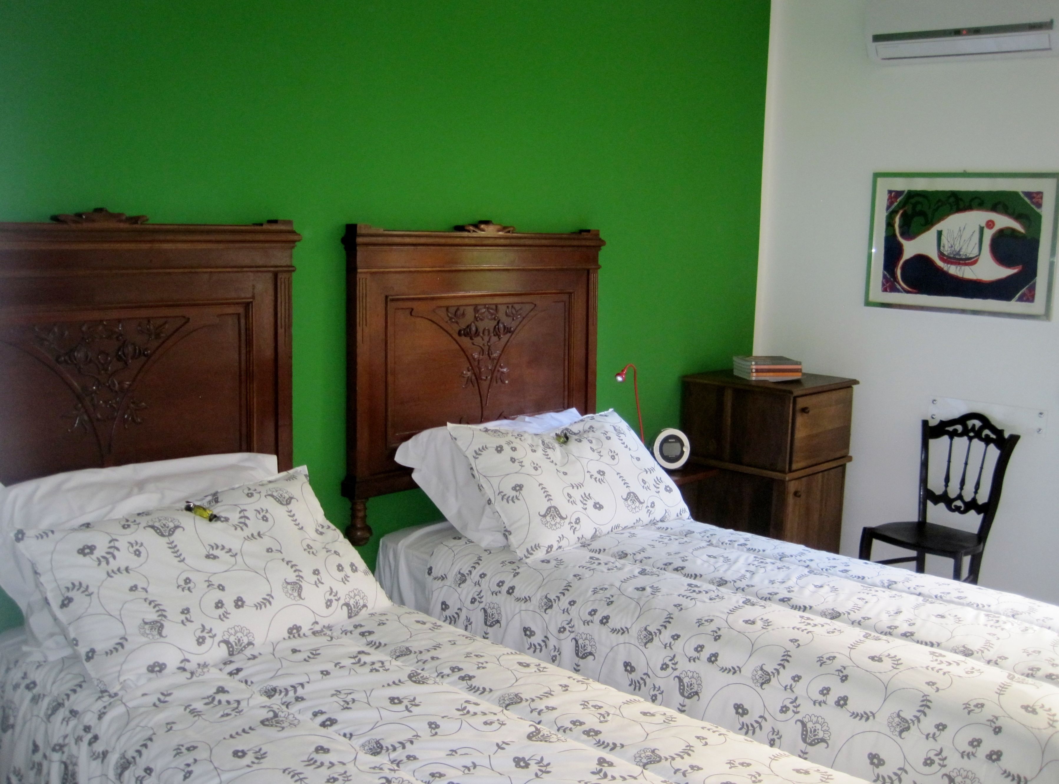 A room with two wooden beds with floral bedding, a small bedside table with a lamp, a chair, and a framed picture on the wall.