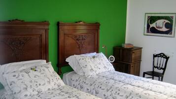 A room with two wooden beds with floral bedding, a small bedside table with a lamp, a chair, and a framed picture on the wall.