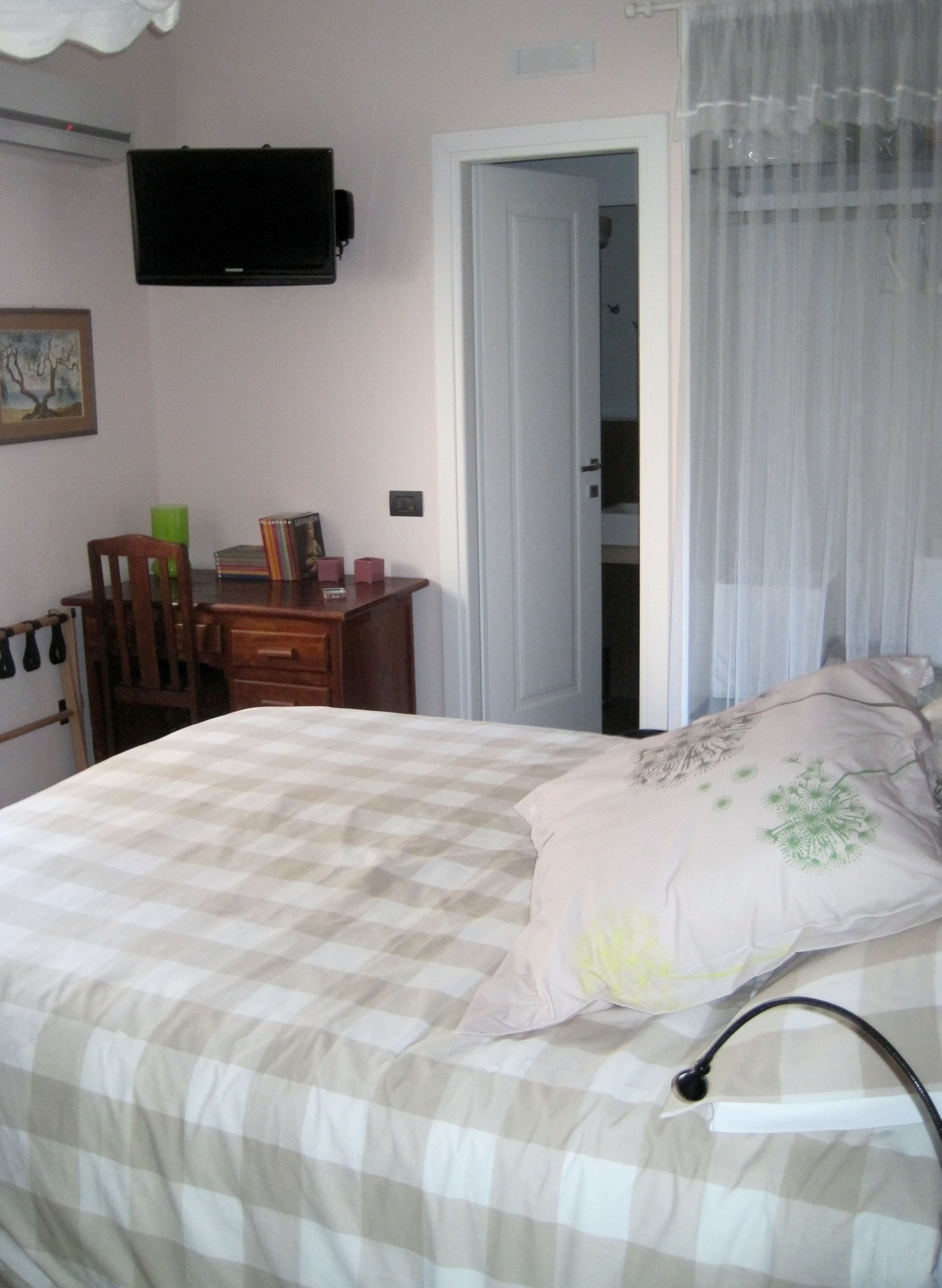 Une chambre avec une couvre-lit à carreaux, une table de chevet avec une lampe, une chaise et un téléviseur fixé au mur.
