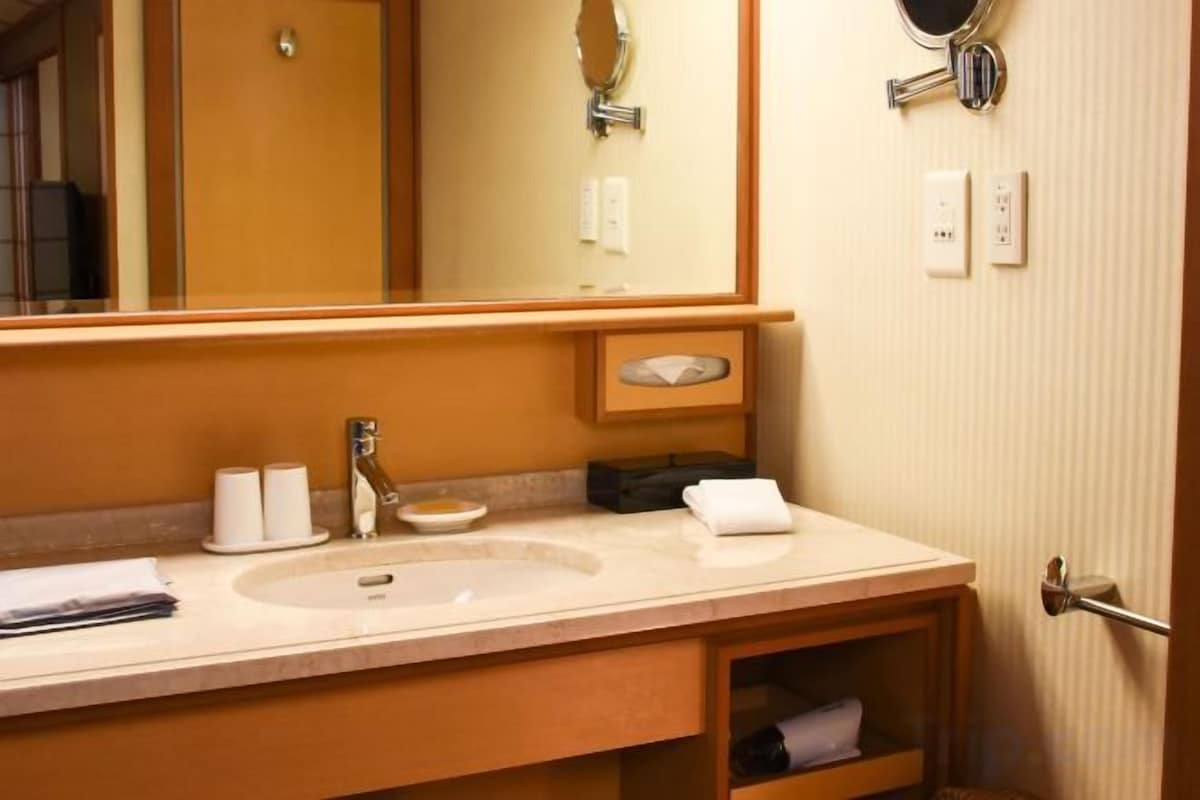 A hotel bathroom with a large mirror, two sinks, a towel rack, and a small shelf with toiletries.