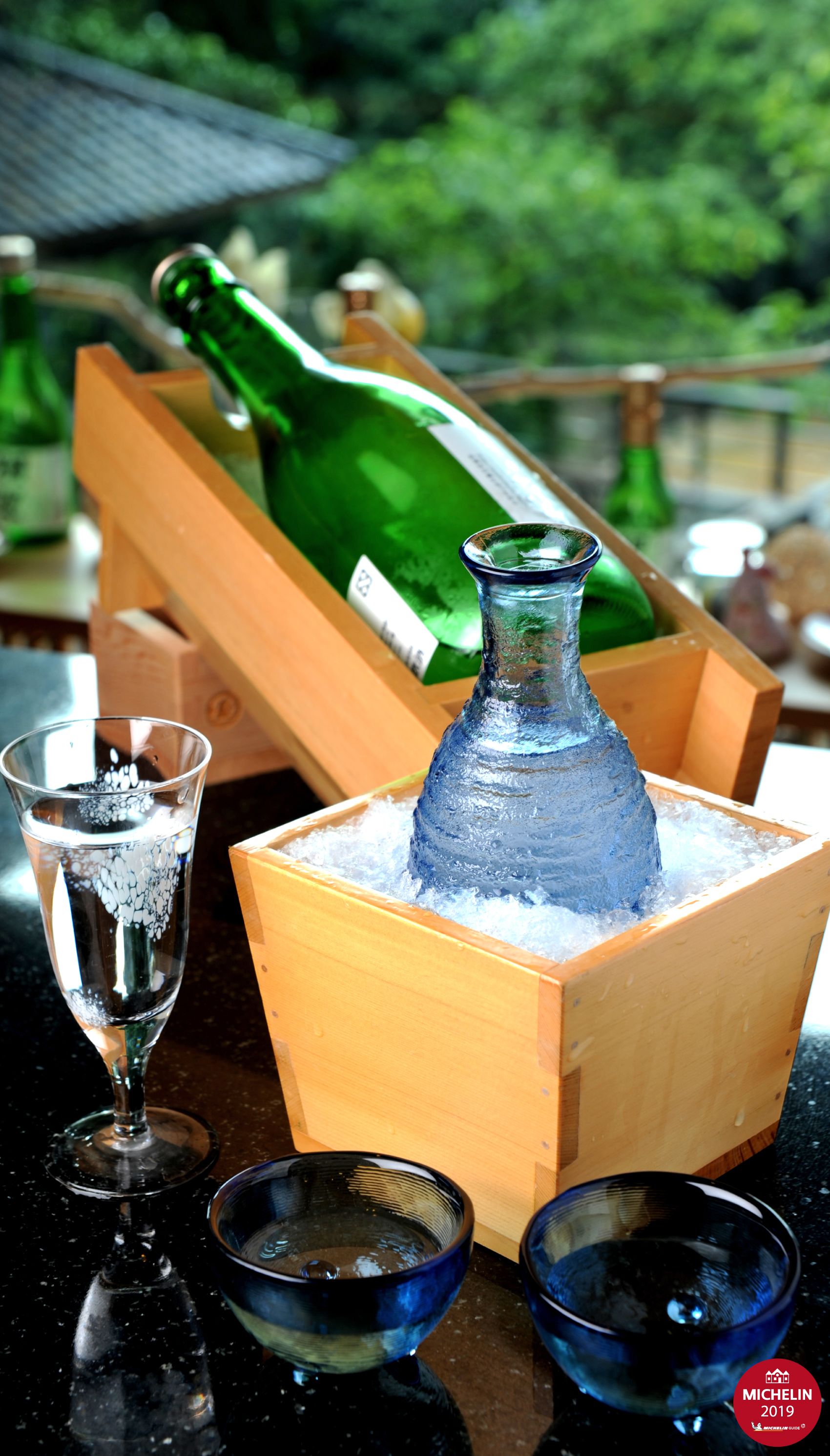 En trälåda som innehåller en grön flaska och en glasflaska, samt ett glas vatten och två skålar som står på ett bord.