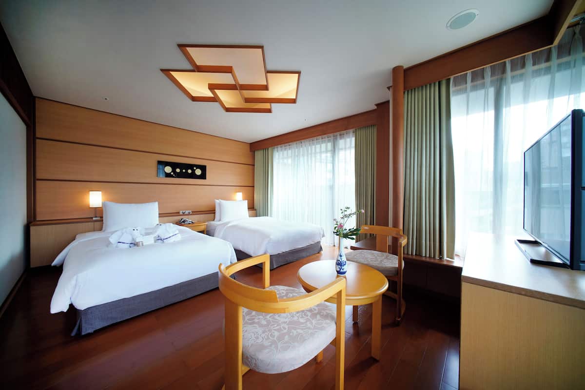 A hotel room with two beds, a desk, a chair, a TV, and a large window with curtains.
