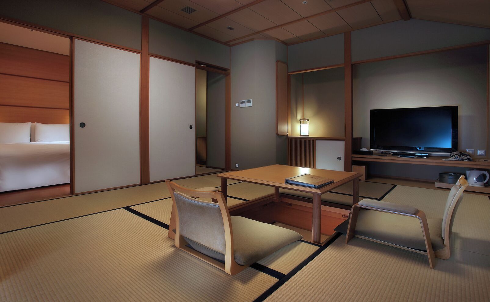 A traditional Japanese-style room with tatami flooring, a low table, two chairs, a bed with sliding doors, a television, and a bedside table with a lamp.