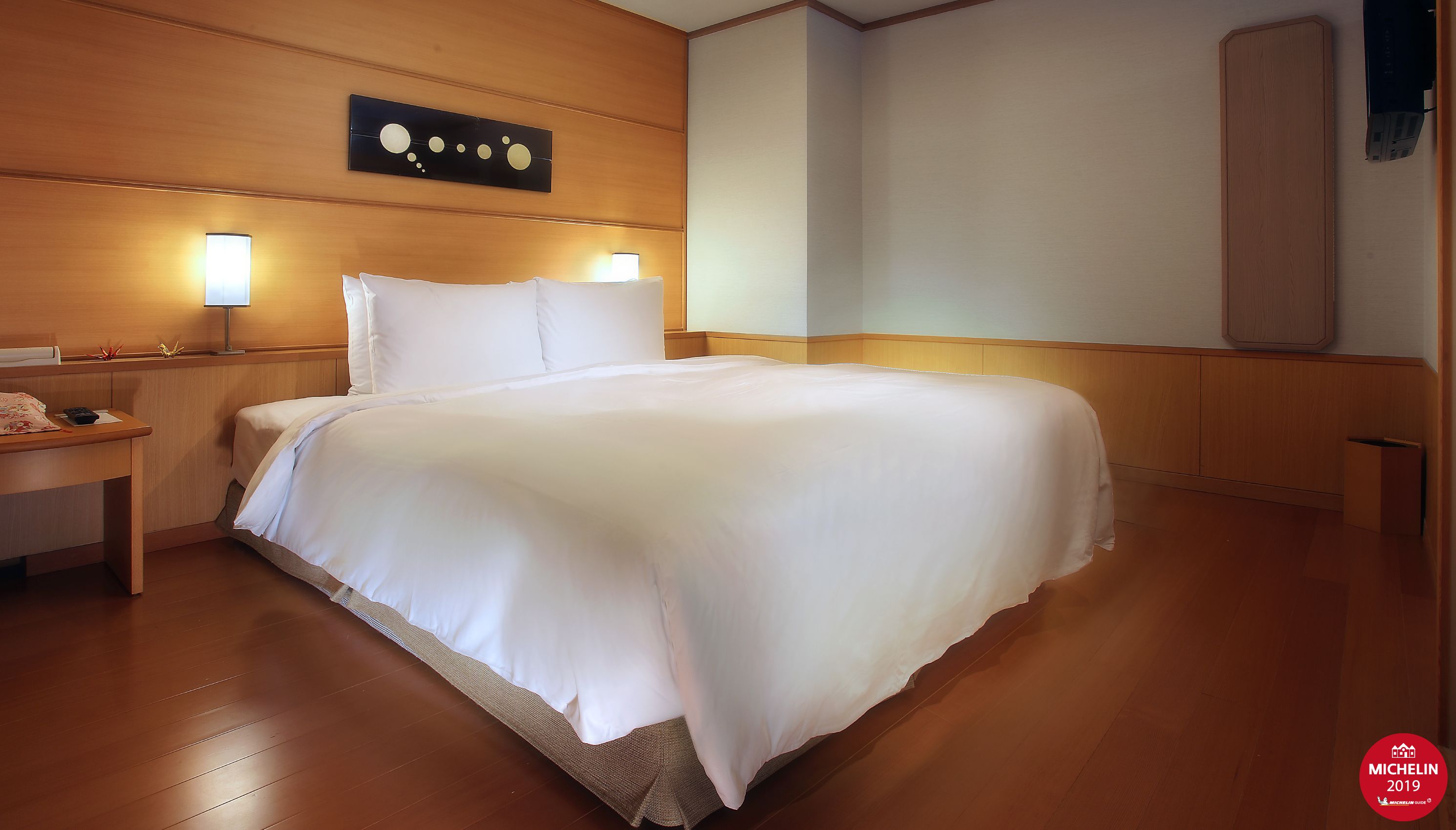 Habitación de hotel con una cama grande, cabecero de madera, lámparas de noche, una mesita de noche de madera y una obra de arte colgada en la pared.