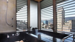 A modern bathroom with a large bathtub, a shower, and a view of the city through large windows.