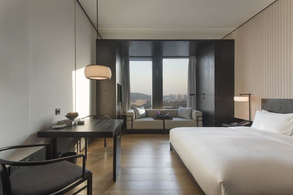 A hotel room with a bed, a desk, a chair, a table, and a large window with a city view.
