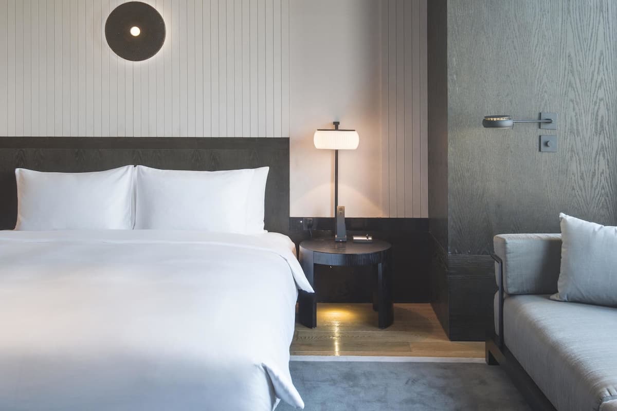 A hotel room with a large bed, a bedside table with a lamp, a sofa, and a wall-mounted light fixture.