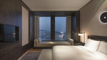 Habitación de hotel con una cama grande, un sofá, un televisor, una mesita de noche, una lámpara y vistas a la ciudad a través de amplias ventanas.