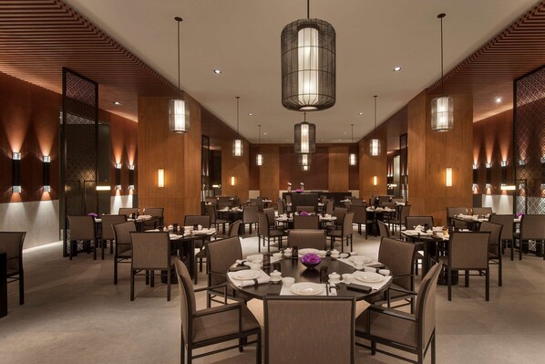 Un vaste espace dédié à la salle à manger, avec de nombreuses tables et chaises, un lustre central et un éclairage chaleureux.