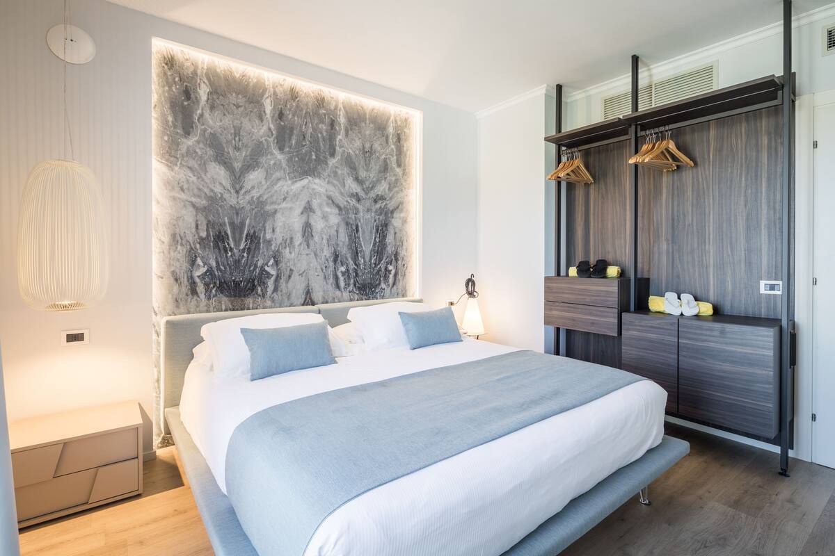 A modern bedroom with a large bed, a wall-mounted artwork, a wardrobe with hanging clothes, and a bedside table with a lamp.