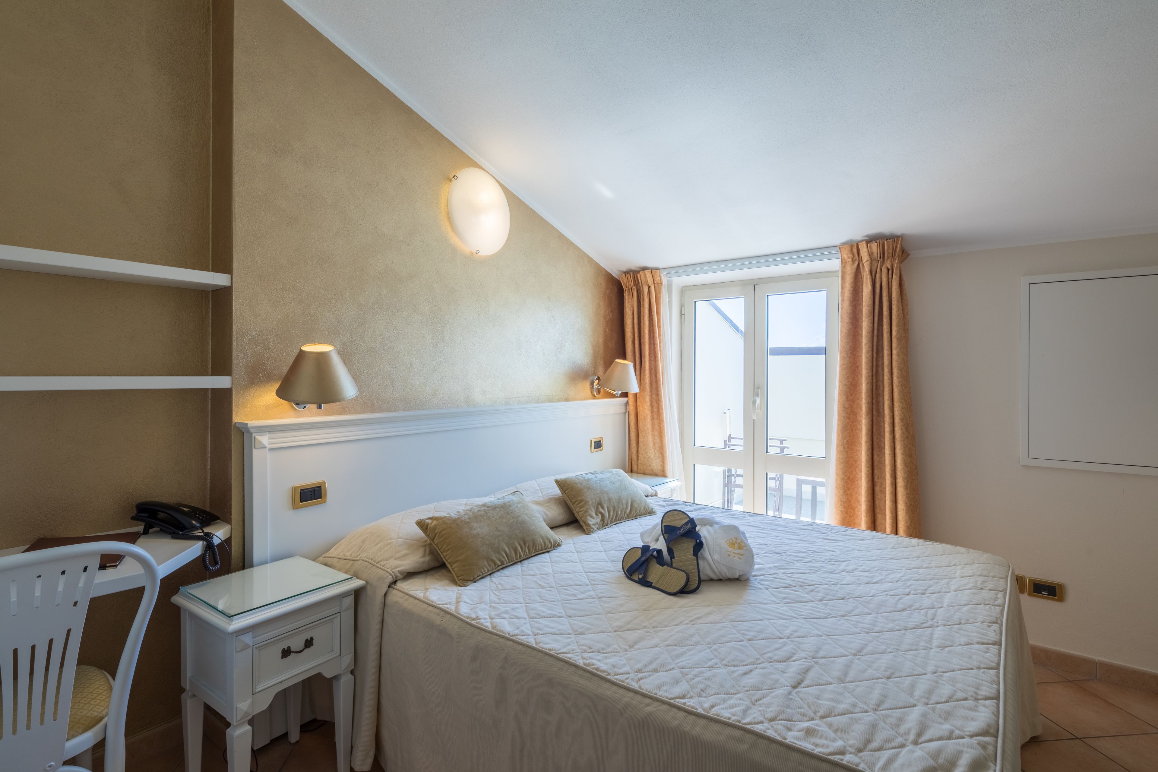 A hotel room with a bed, bedside tables, a desk, a chair, and a window with curtains.