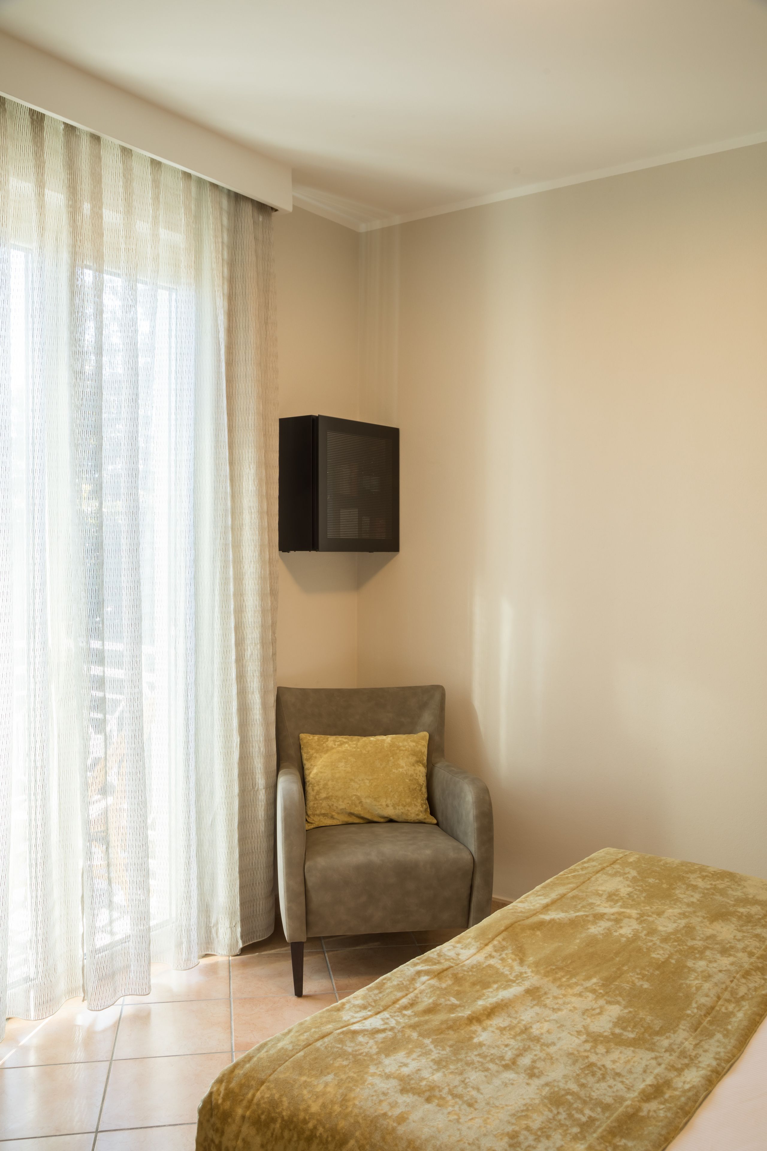 A room with a bed, a chair, a television mounted on the wall, and a window with sheer curtains.