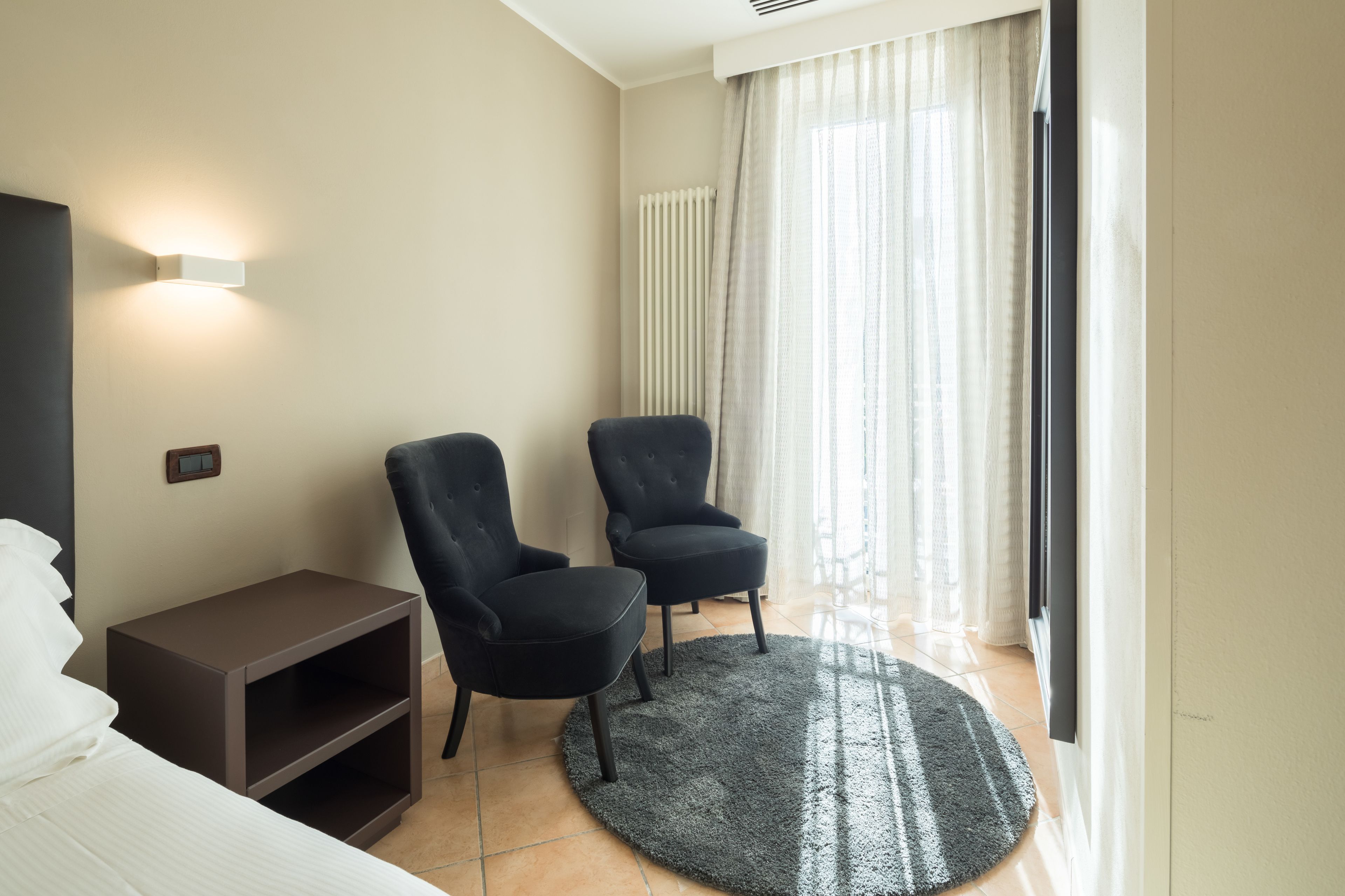 Una habitación de hotel con dos sillones negros, una mesita de noche marrón pequeña, una cama blanca y un ventanal grande con cortinas translúcidas.