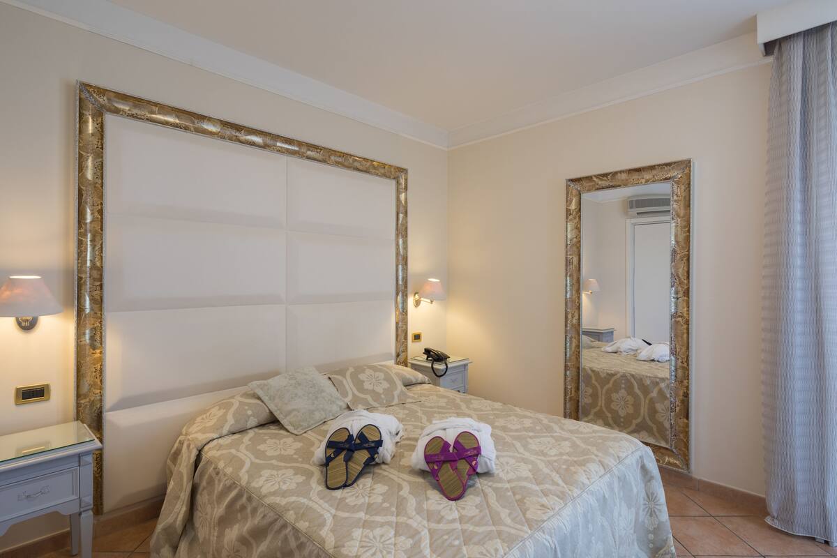 A hotel room with a large bed, a bedside table with a lamp, a nightstand with a phone, and a mirror on the wall.