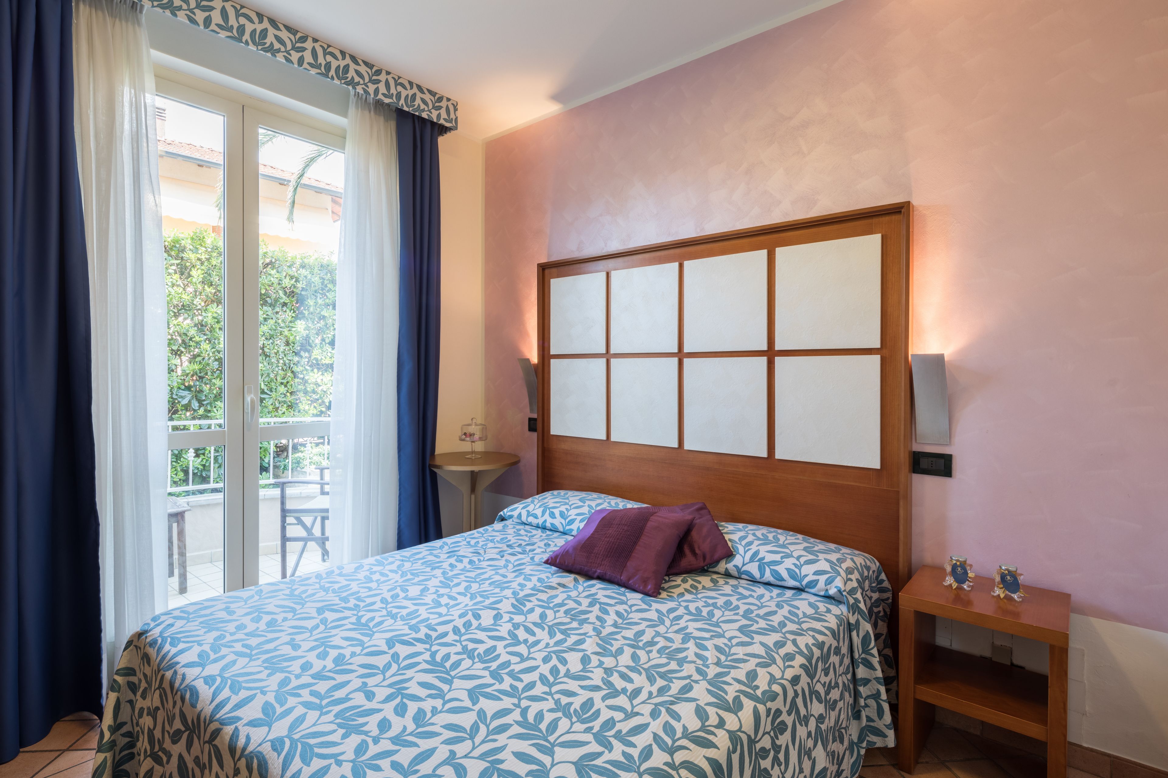 Ein Schlafzimmer mit einem großen Bett, einem Holzkopfende, einem Nachttisch und Blick auf einen Balkon mit Tisch und Stühlen.