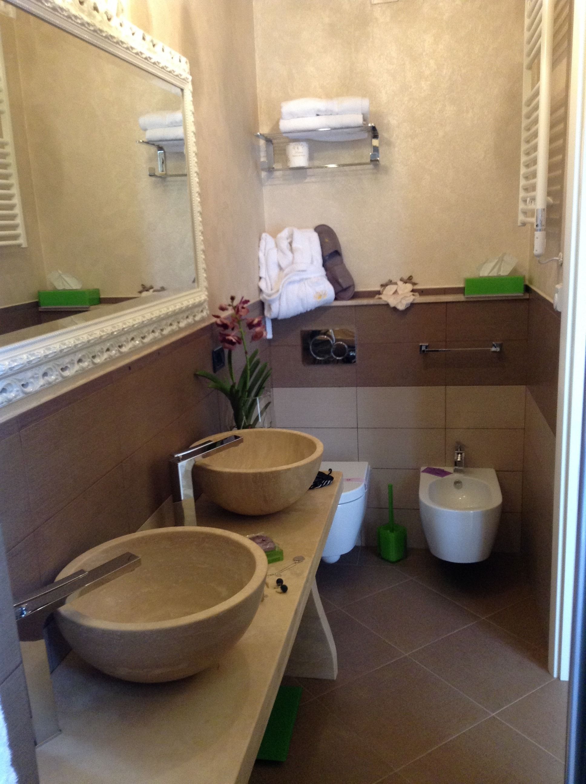 Casa de banho com duas grandes pias de madeira, sanita e bidé. Espelho com moldura decorativa, porta-toalhas e uma pequena planta.