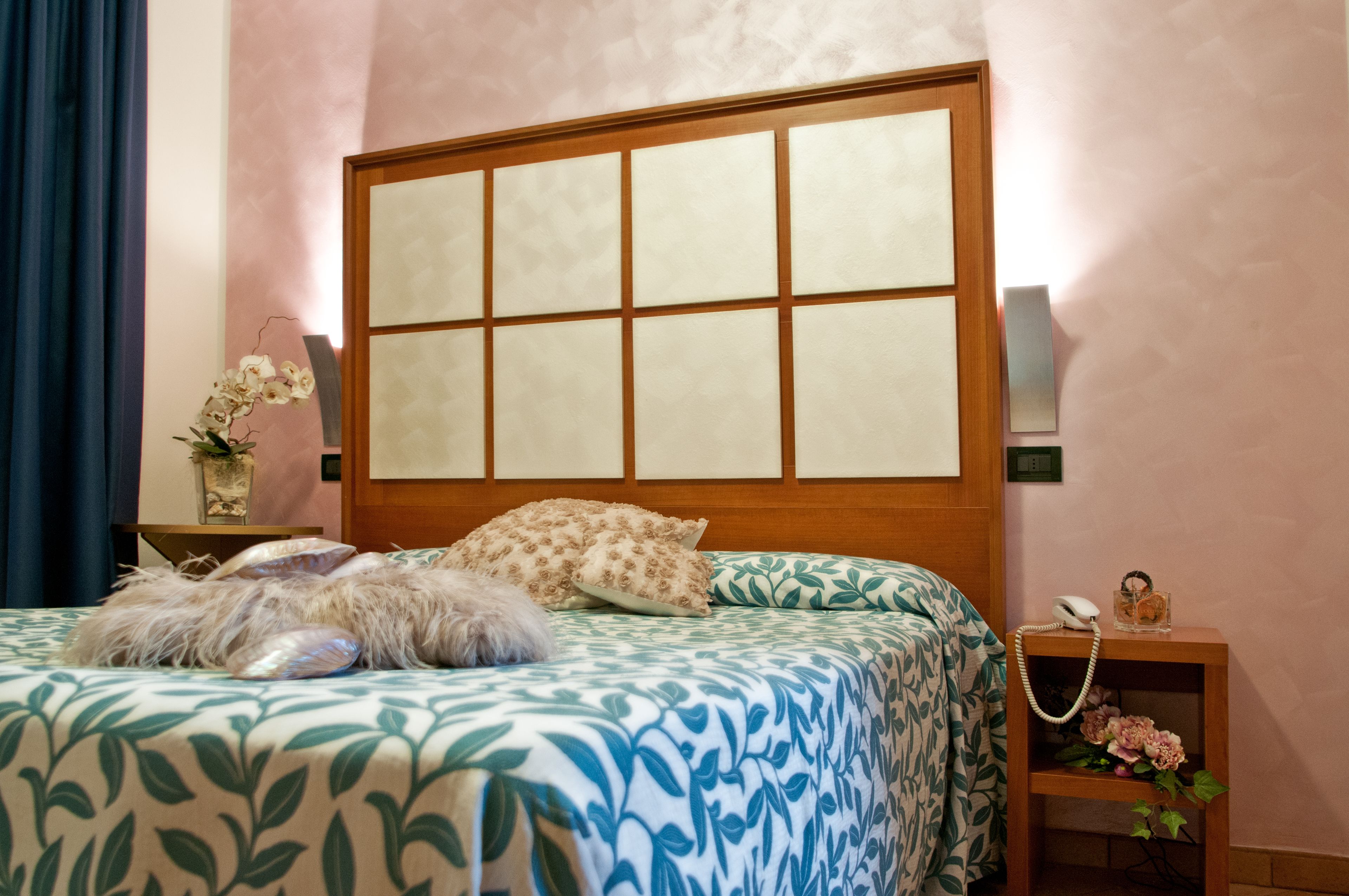 A bedroom with a large bed, a wooden headboard, a bedside table with a lamp, and a small shelf with a phone and decorative items.