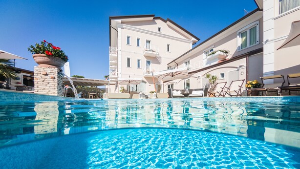A swimming pool with clear blue water, surrounded by a building with balconies and outdoor seating. - Hotel Luna (Massa)