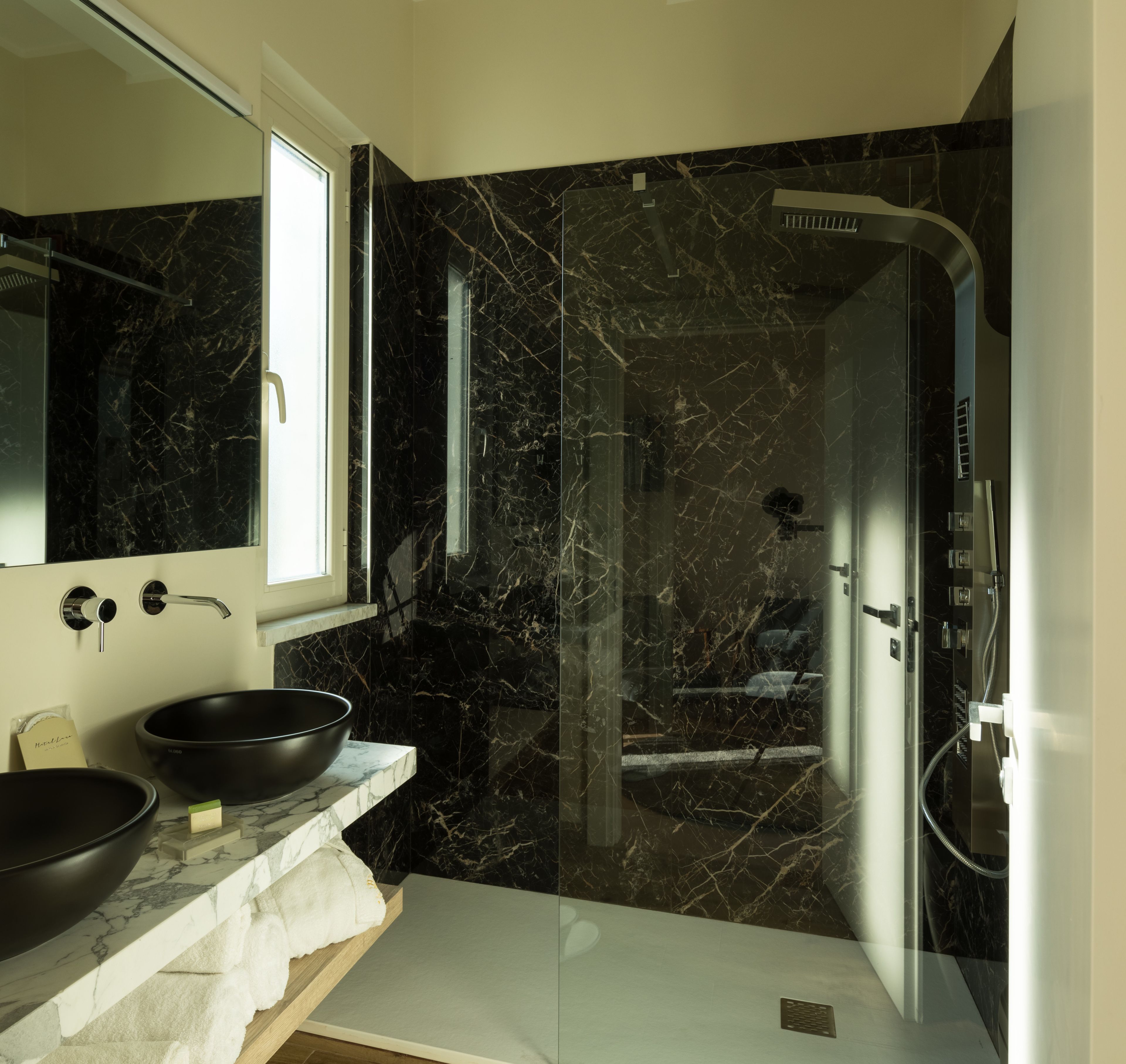 A modern bathroom with a black marble wall, a large mirror, a black basin, and a glass-enclosed shower area.