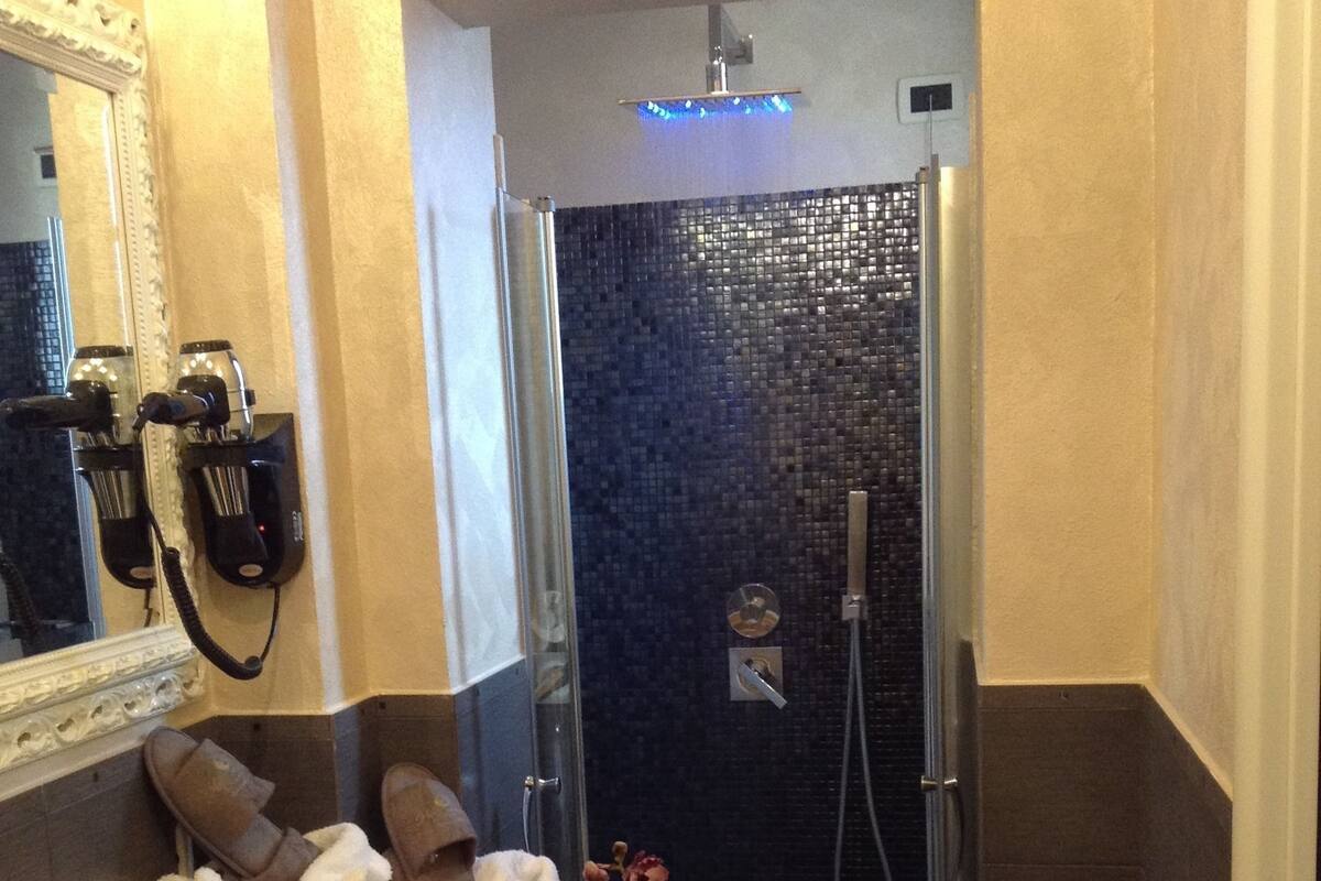 A bathroom with a wooden sink, a mirror, a towel rack, a shower with a dark tiled wall, and a vase with flowers.