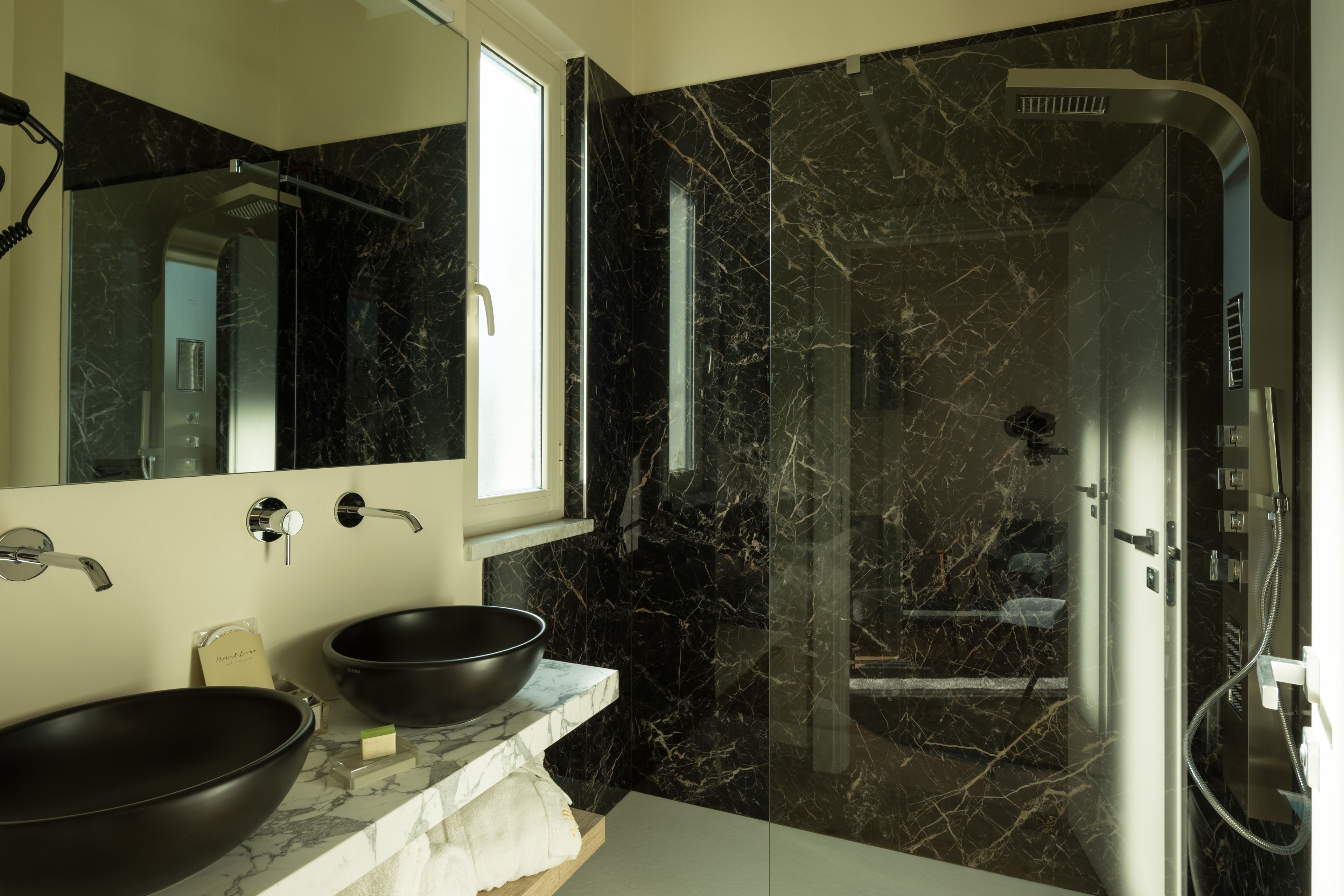 Un bagno moderno con un grande specchio, due lavabi a incasso neri, una cabina doccia in vetro e pareti in marmo.
