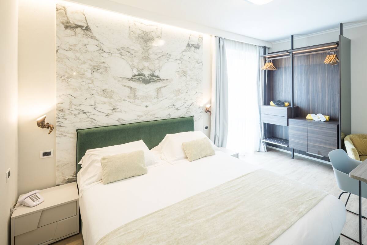 A modern hotel room with a marble headboard, a bed with white linens, a bedside table with a phone, and a seating area with a chair and a small table.