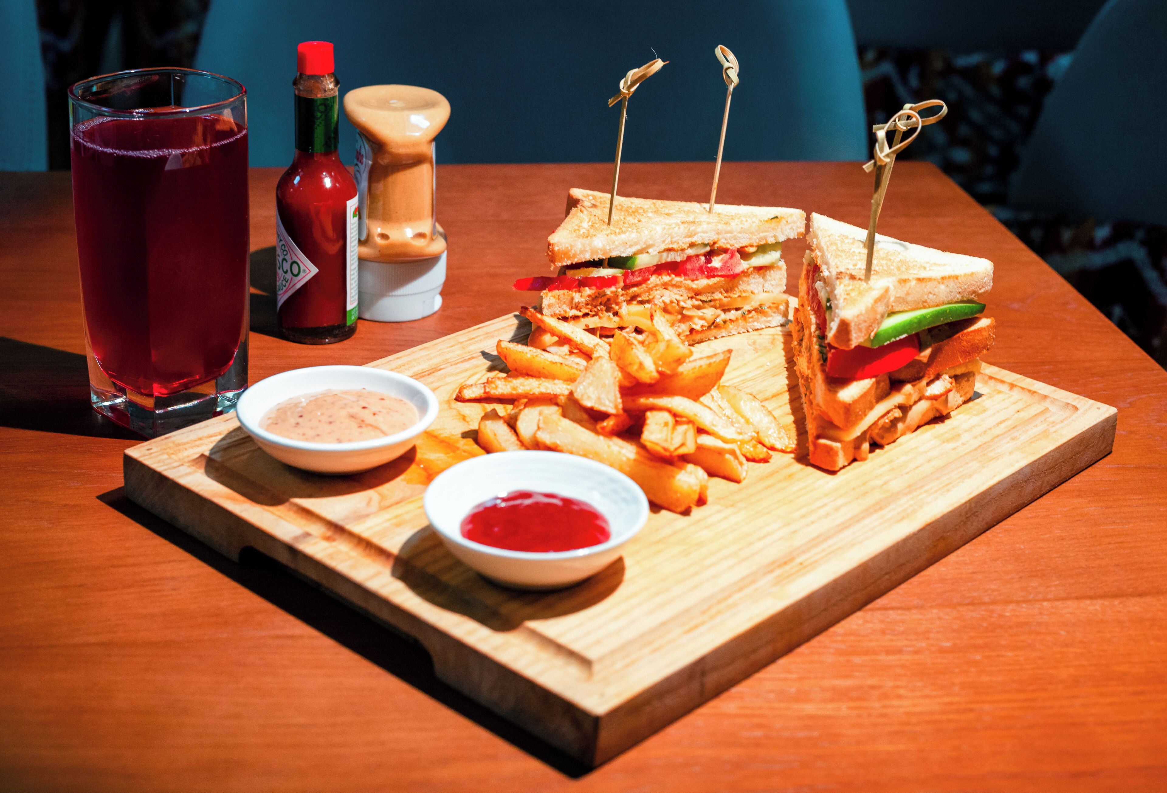 A wooden table with a sandwich cut in half, a plate of fries, a glass of red drink, and two small bowls of condiments.