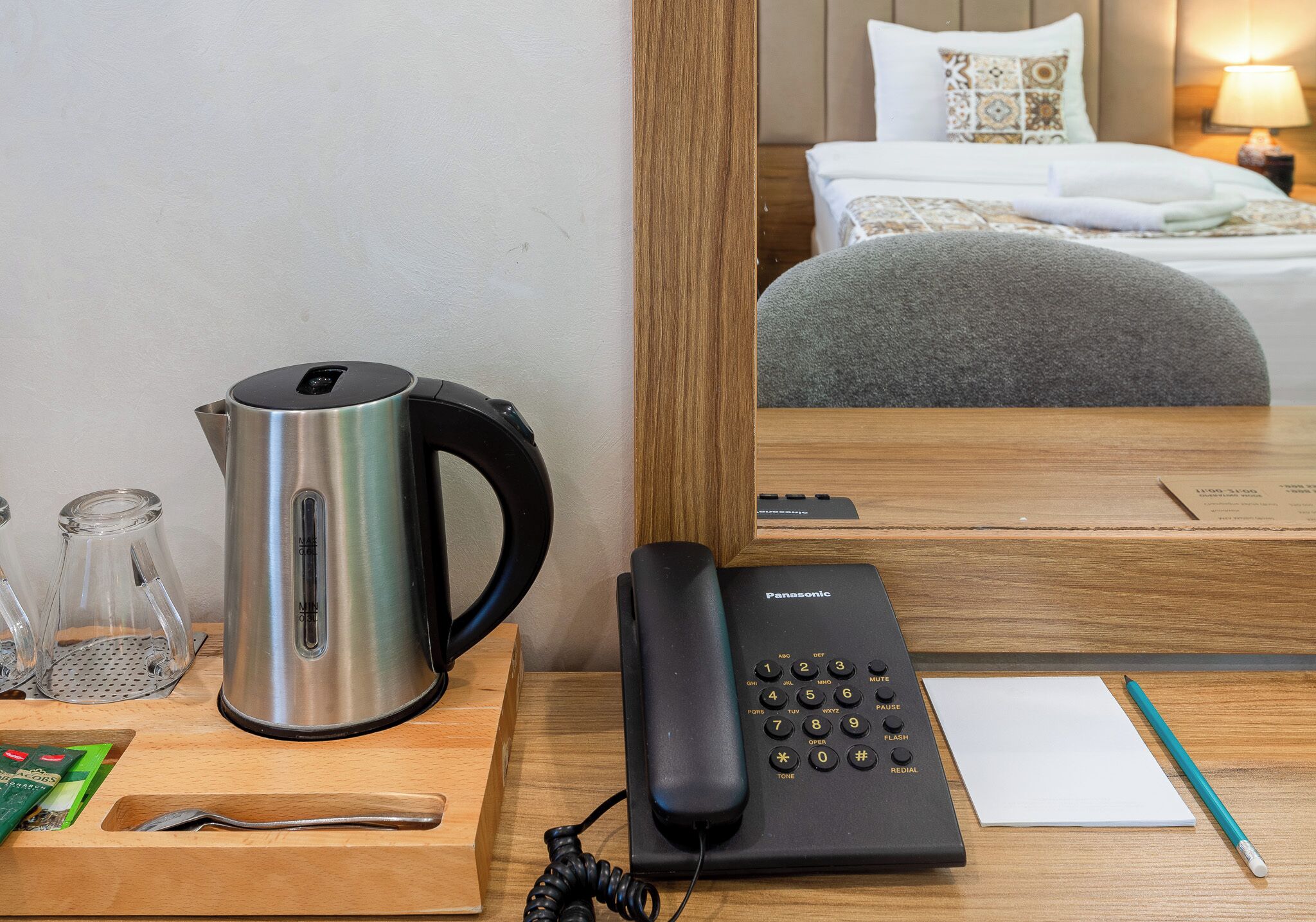 Habitación de hotel con cama, mesita de noche con tetera, vasos, teléfono y bloc de notas.