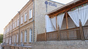 A historic building with a sign reading "Malika Hotel". It features a brick facade, large windows, and a wooden balcony with curtains.