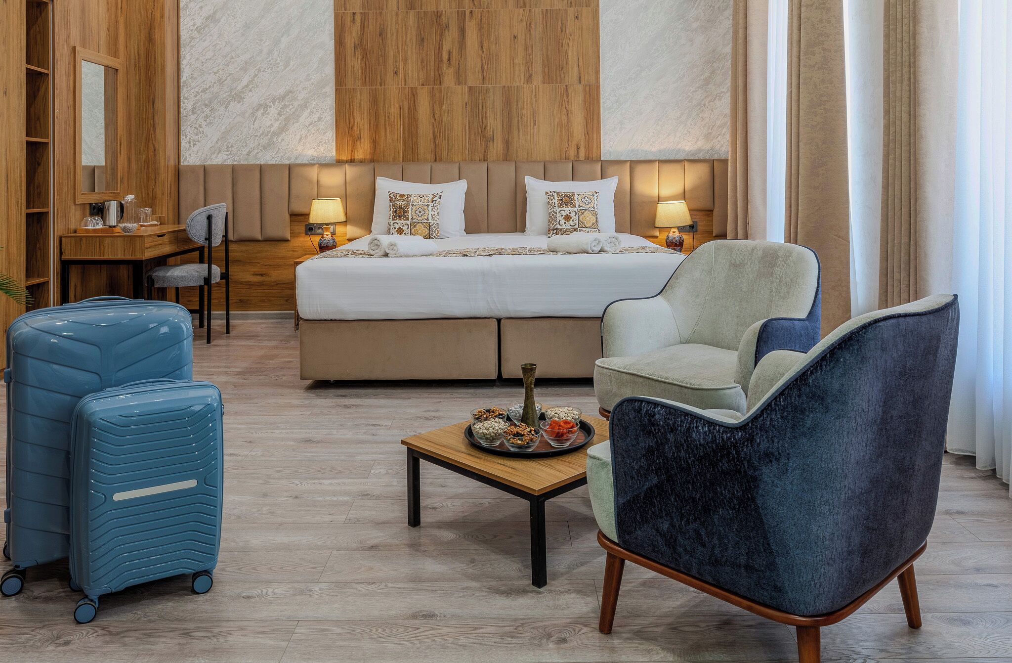 A hotel room with a bed, two armchairs, a small table, and a desk with a chair. There are two suitcases and a vase on the table.