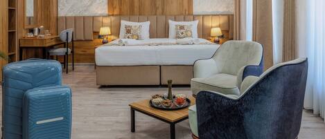 A hotel room with a bed, two armchairs, a small table, and a desk with a chair. There are two suitcases and a vase on the table.