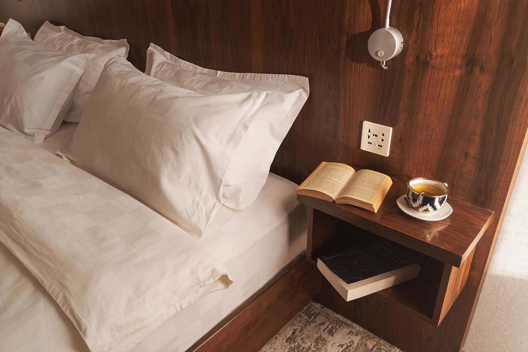 A neatly made bed with white linens, a bedside table with a book and a cup of tea, and a wall socket.