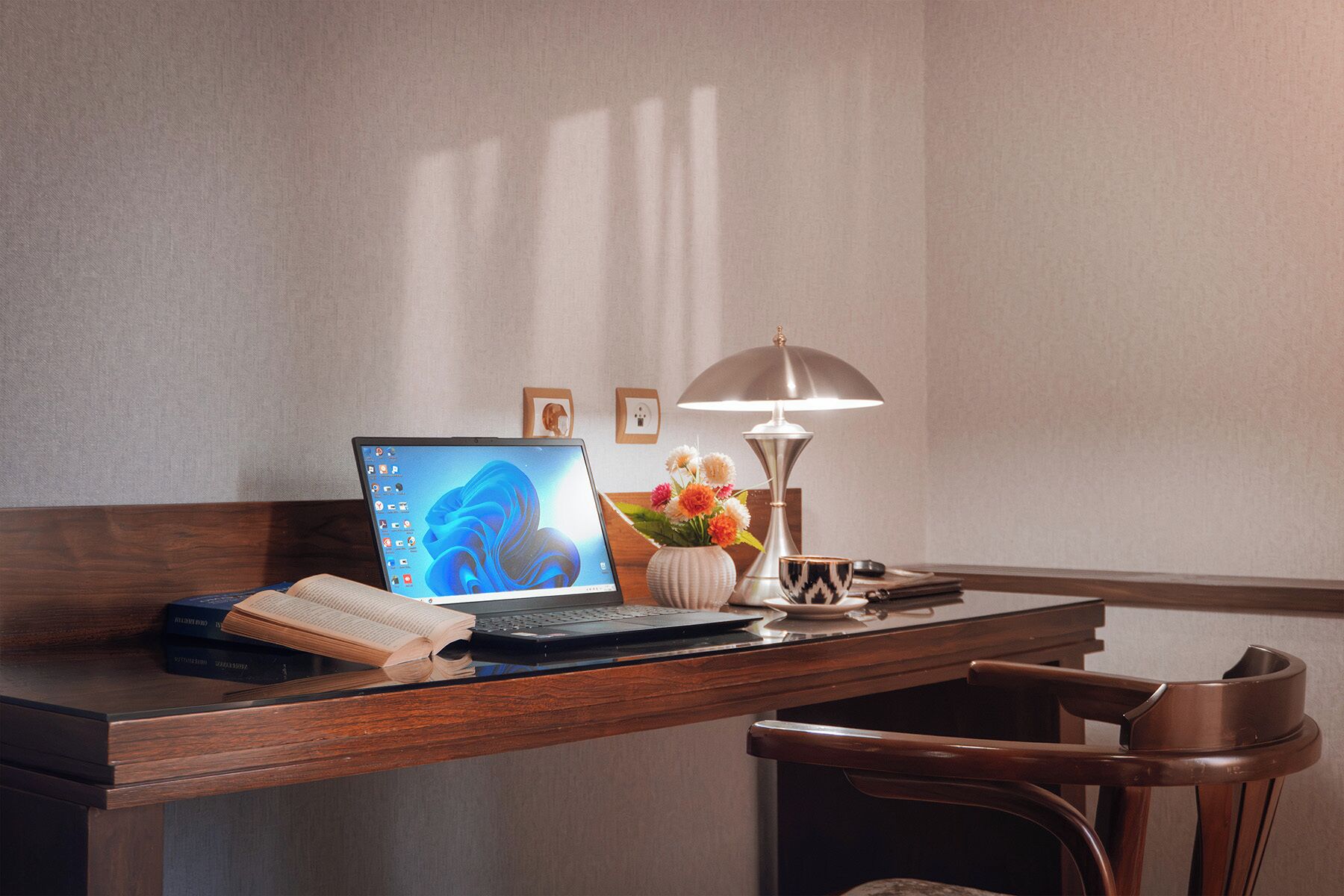 A wooden desk with a laptop, a book, a vase with flowers, a lamp, and a chair.