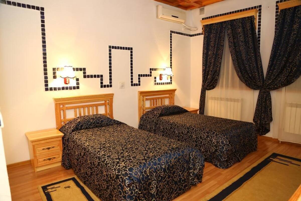 Un dormitorio con dos camas, cabeceros de madera, mesitas de noche, una colcha estampada, una lámpara de pared y una ventana con cortinas.