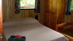 A single bed with a green towel and a pink flower, a wooden door, and a window with curtains. - Vaianae Lodge (Moorea-Maiao)