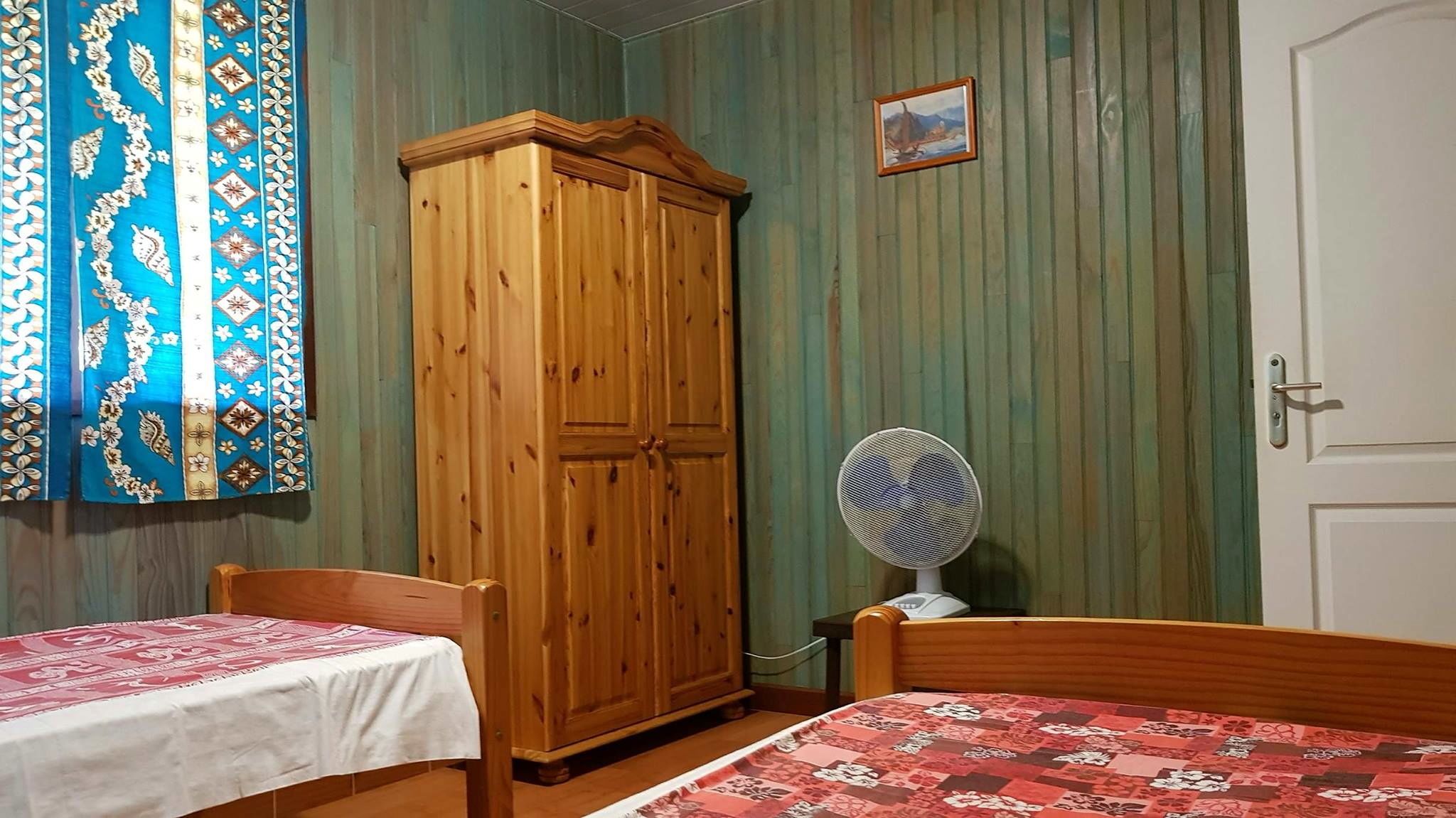 Ein Zimmer mit zwei Holzbetten, einem Holzschrank, einem Tisch mit weißem Tuch und einem Ventilator, sowie einem gemusterten Vorhang.