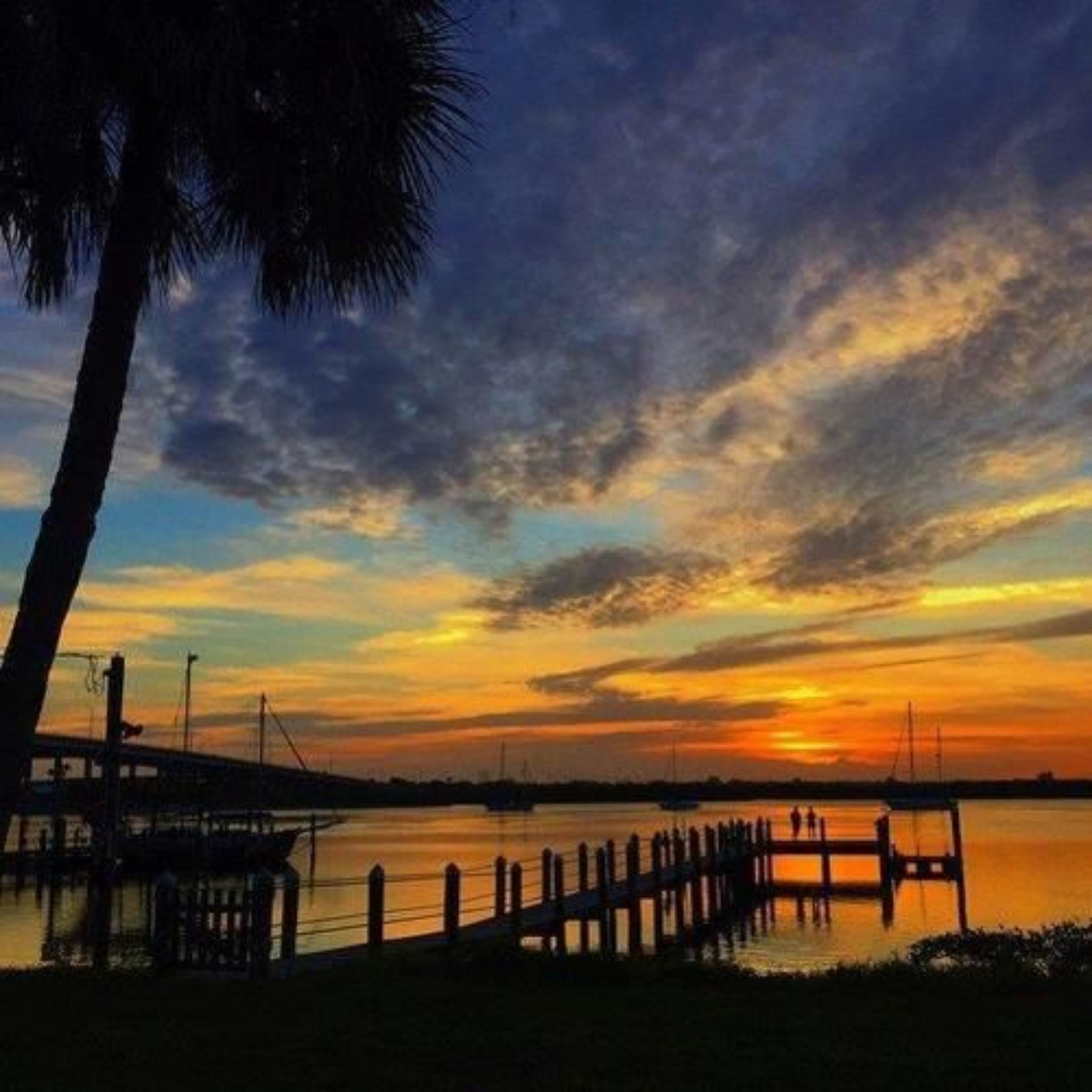 Ein Sonnenuntergangsblick von einer Anlegestelle mit einer hölzernen Pier, die sich in ruhiges Wasser erstreckt, umgeben von einem klaren Himmel mit vereinzelten Wolken.