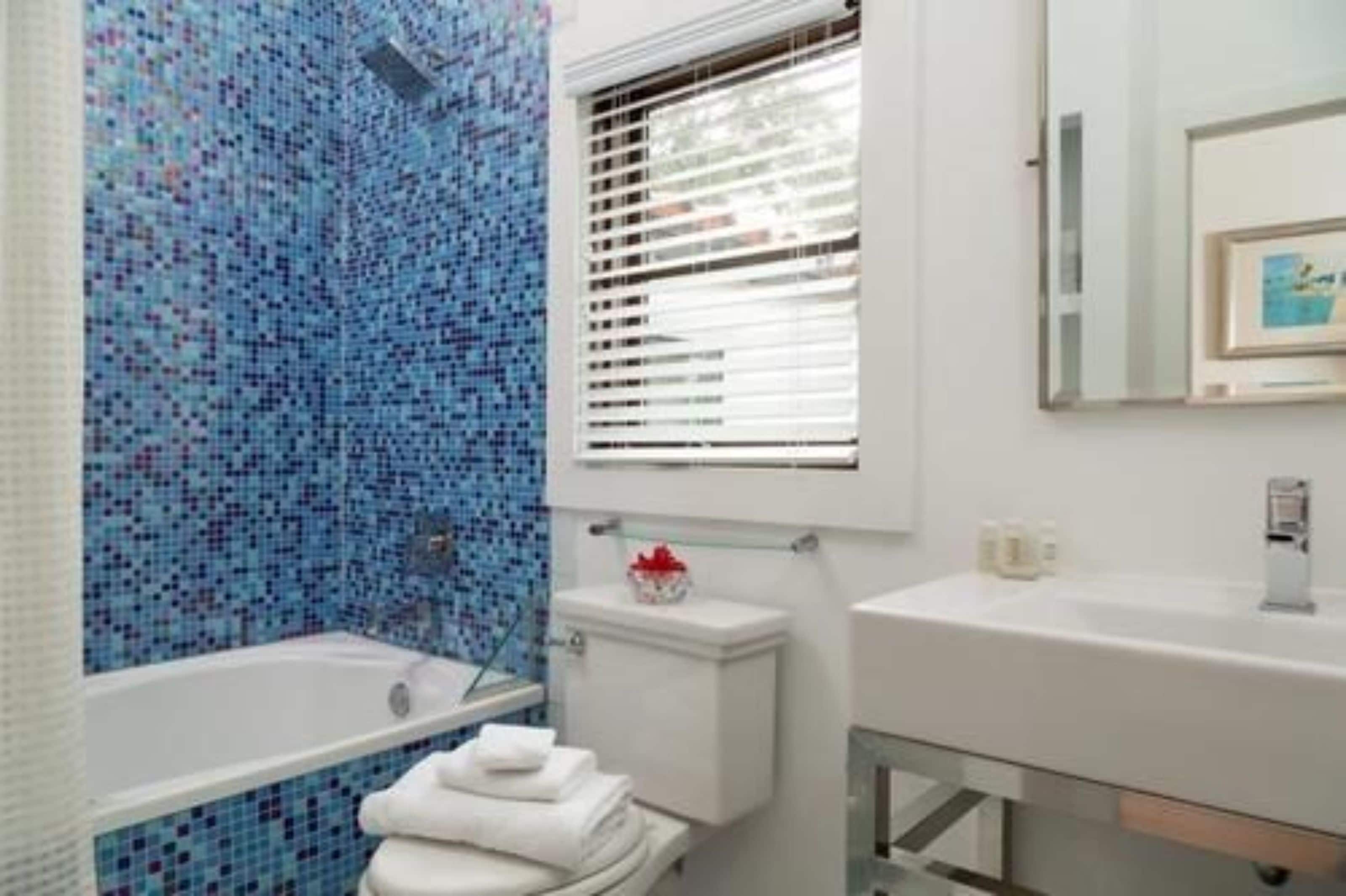 Ein Badezimmer mit Wand aus Mosaikfliesen, einer Badewanne, einer Toilette, einem Waschbecken und einem Fenster mit Jalousien.