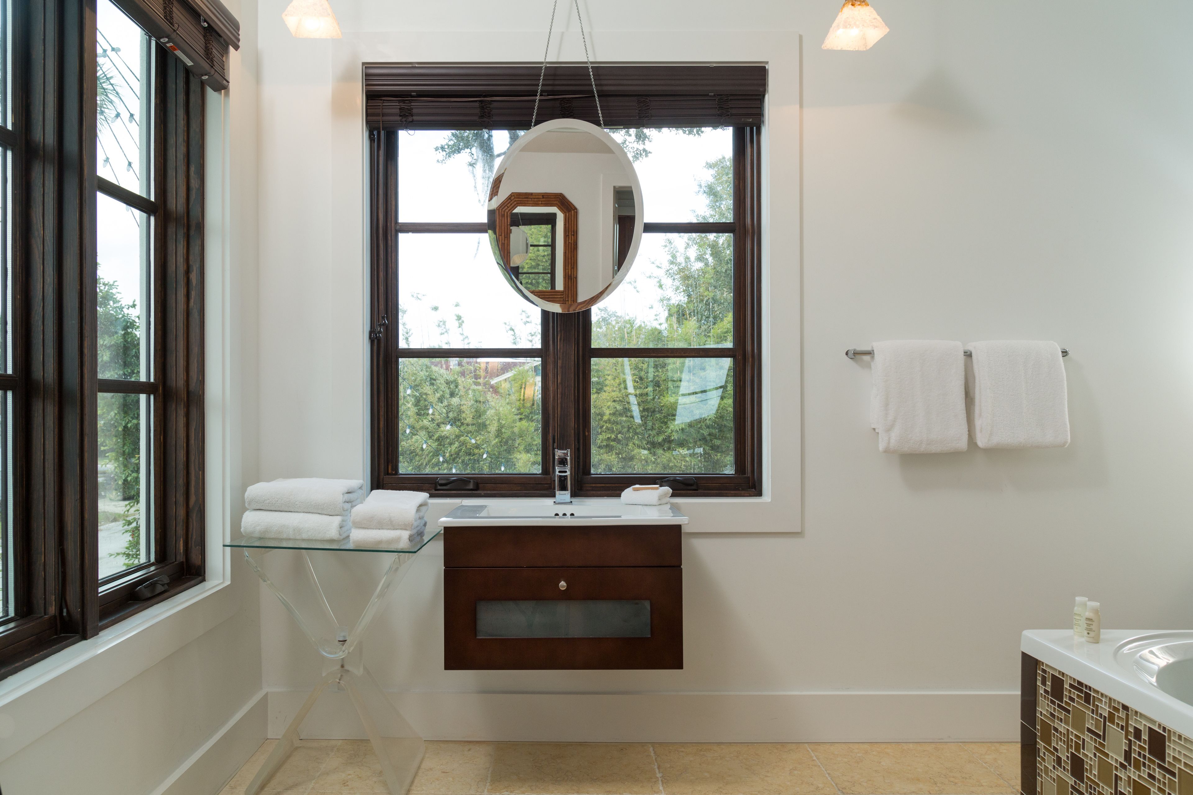 Un baño con estante de vidrio, espejo redondo, ventana y bañera.