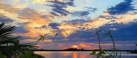 A sunset over a calm lake with a distant shoreline and some vegetation in the foreground.