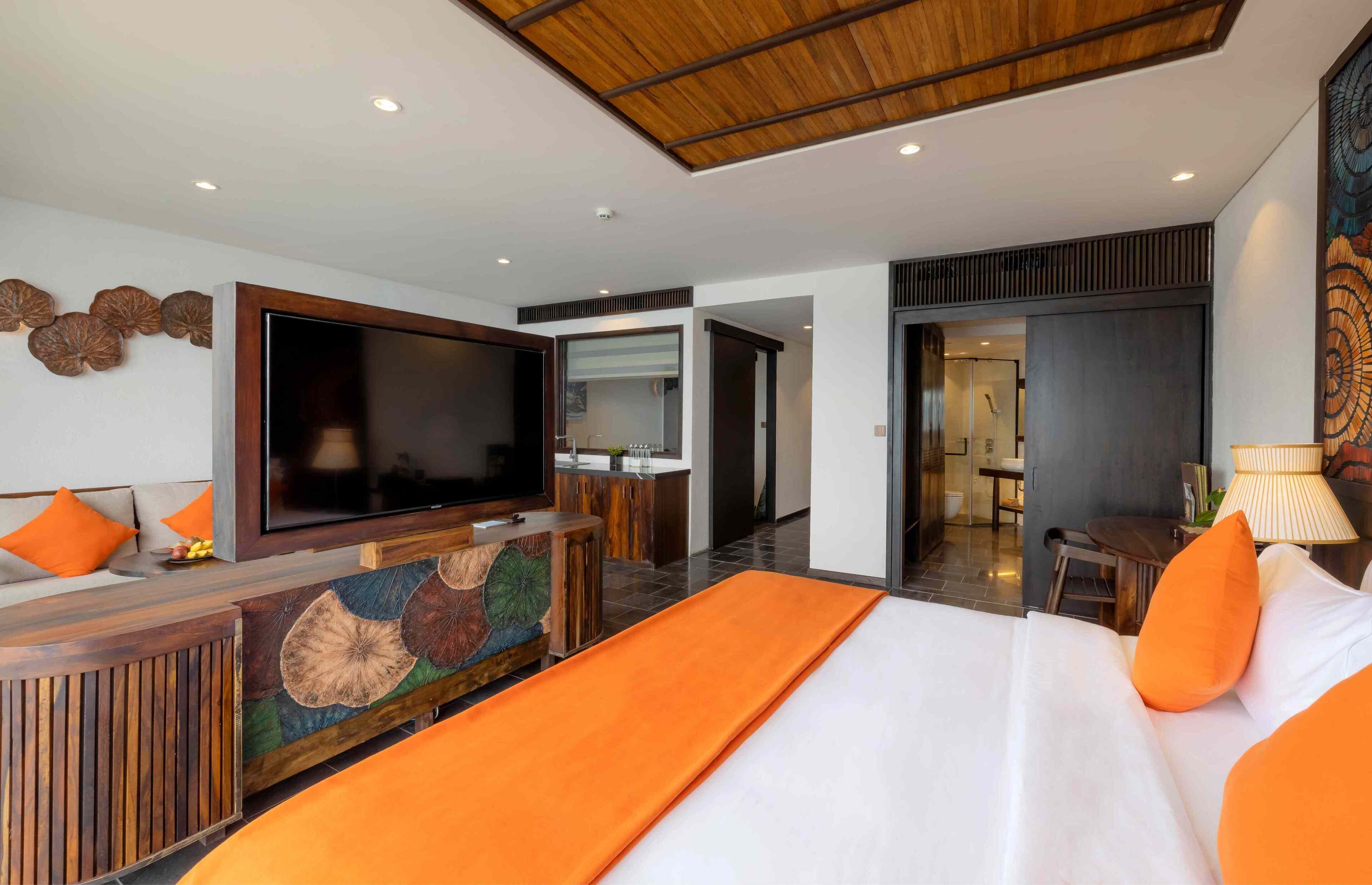 Habitación de hotel moderna con un televisor de pantalla plana grande, un cabecero de madera, una pared de acento naranja y vistas al baño.